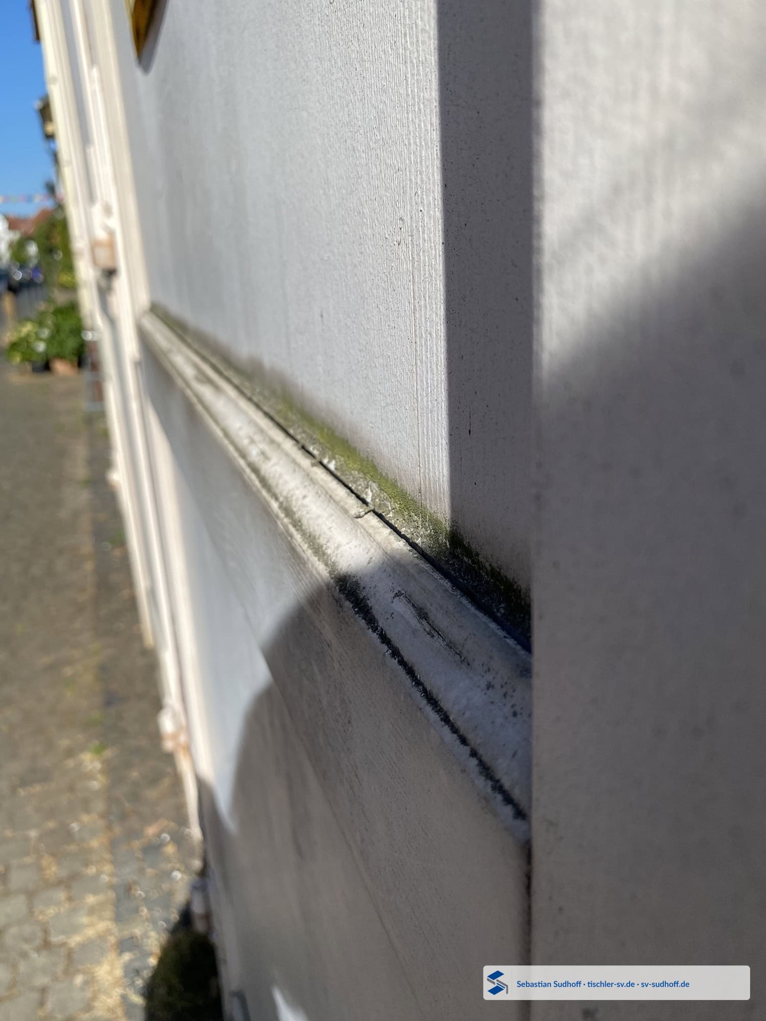 Abgelöste Silikonfuge an der Holzfüllung eines weißen Garagentors mit grünlichem Bewuchs